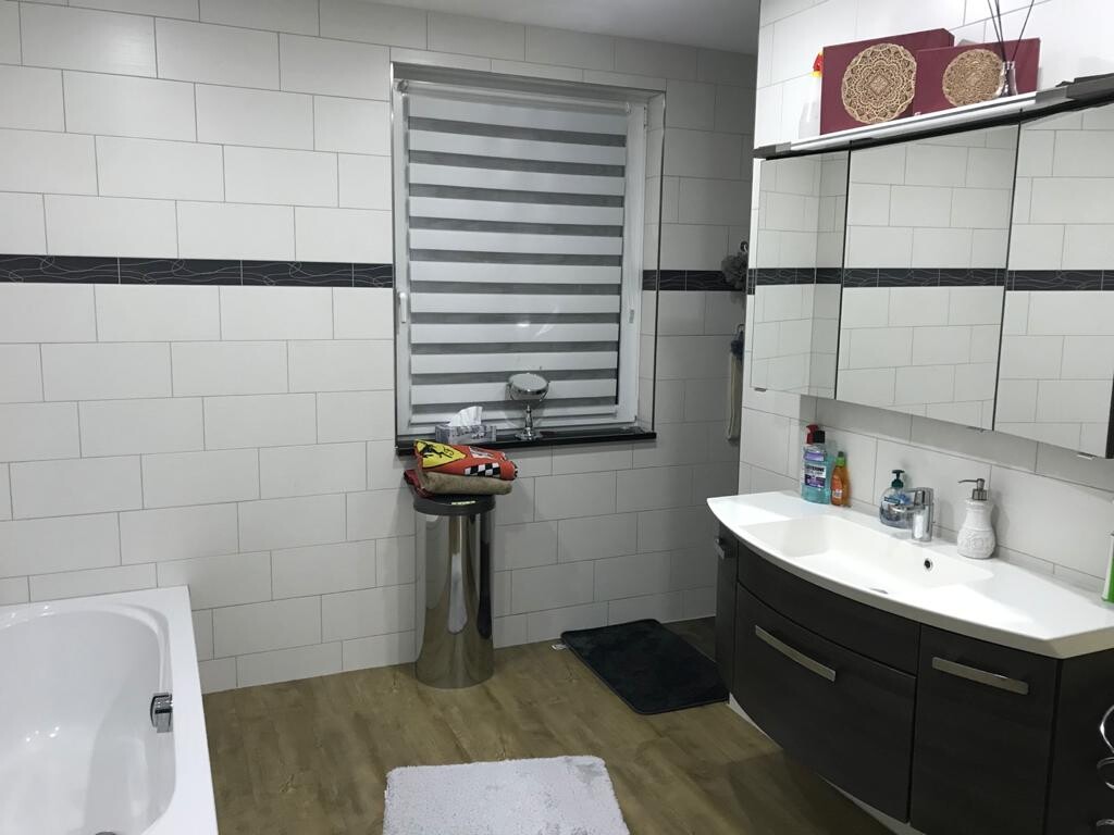 Modernes Badezimmer mit weißen Fliesen, Badewanne links, Waschbecken, Spiegel und Fenster mit Rollo