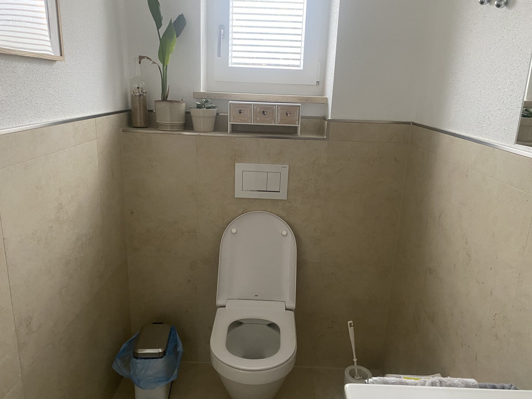 Badezimmer mit WC, Spülplatte, beigen Fliesen, Fenster über der Toilette, Pflanzendeko.