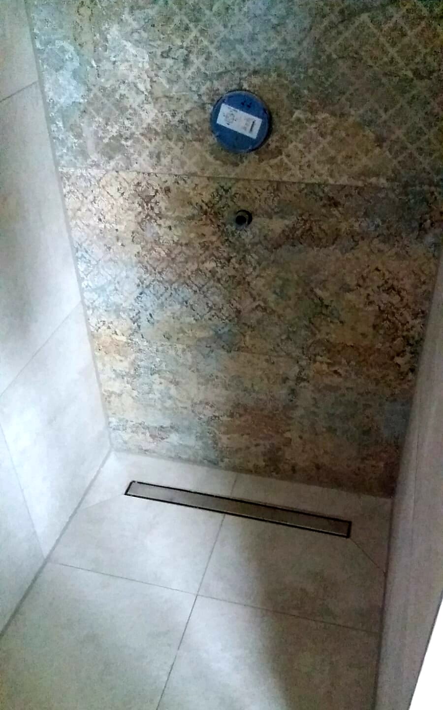 Dusche mit gemusterten Wandfliesen, blauem runden Ventildeckel an der Wand und Bodenablauf.