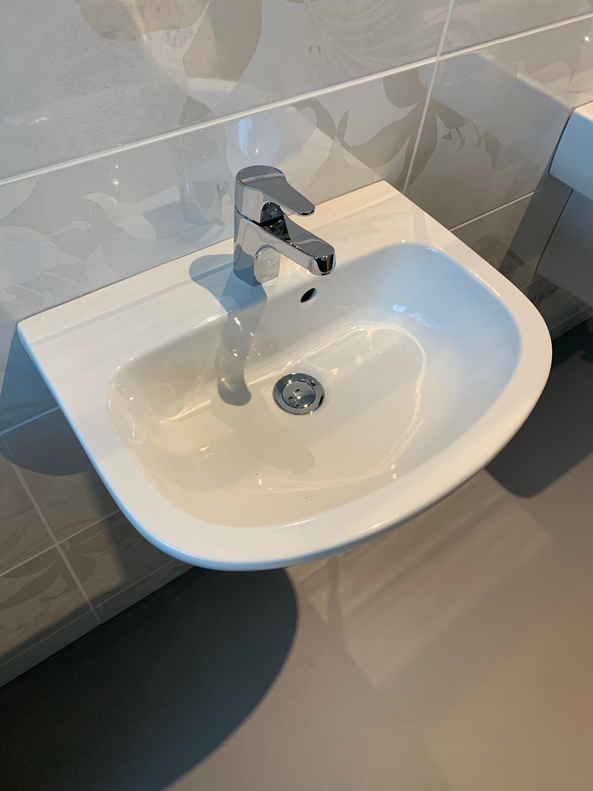 Weißes Waschbecken mit silberner Einhebelarmatur vor Fliesen mit Blättermuster.