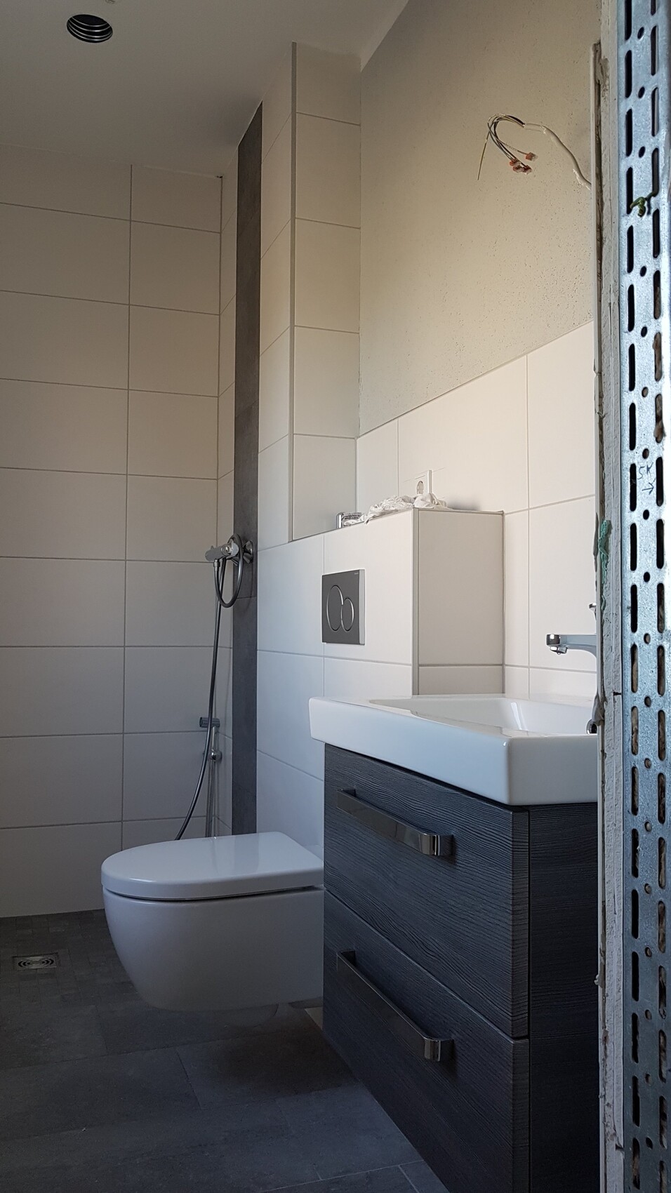 Badezimmer mit Wand-WC, Waschbecken auf dunklem Holzunterschrank, Dusche links, freiliegende Kabel.