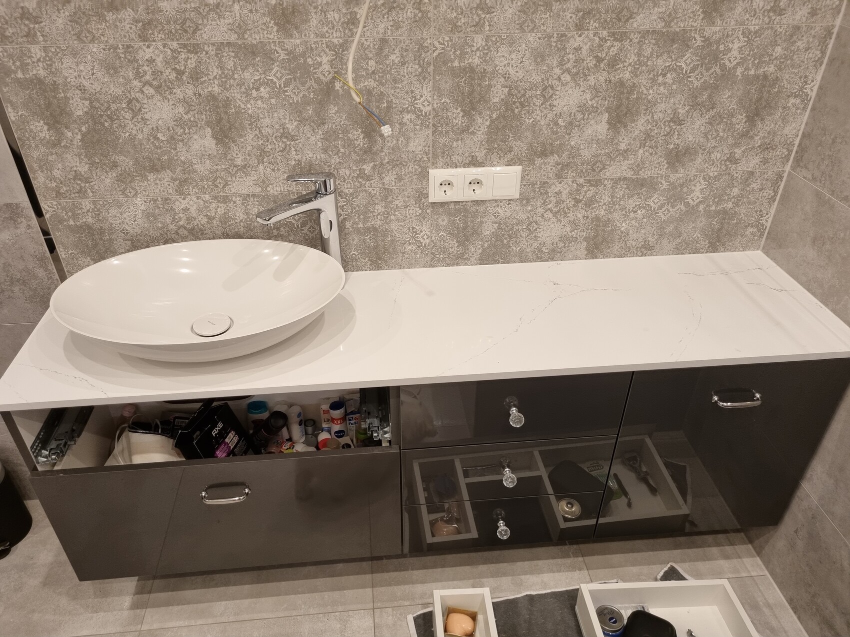 Moderne Waschtischplatte mit rundem Becken, Chromarmatur, graue Fliesenwand, offene Schränke.