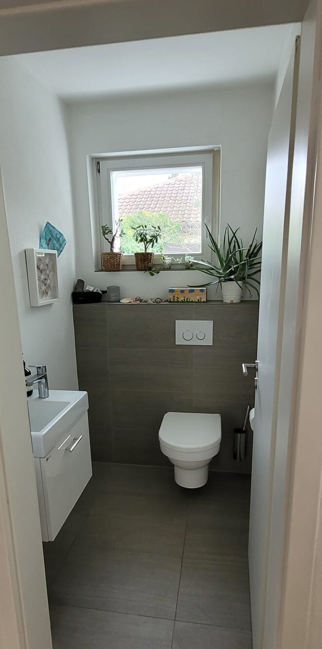 Kleines Badezimmer mit Wandtoilette, Waschbecken-Unterschrank links, Fenster mit Pflanzen.