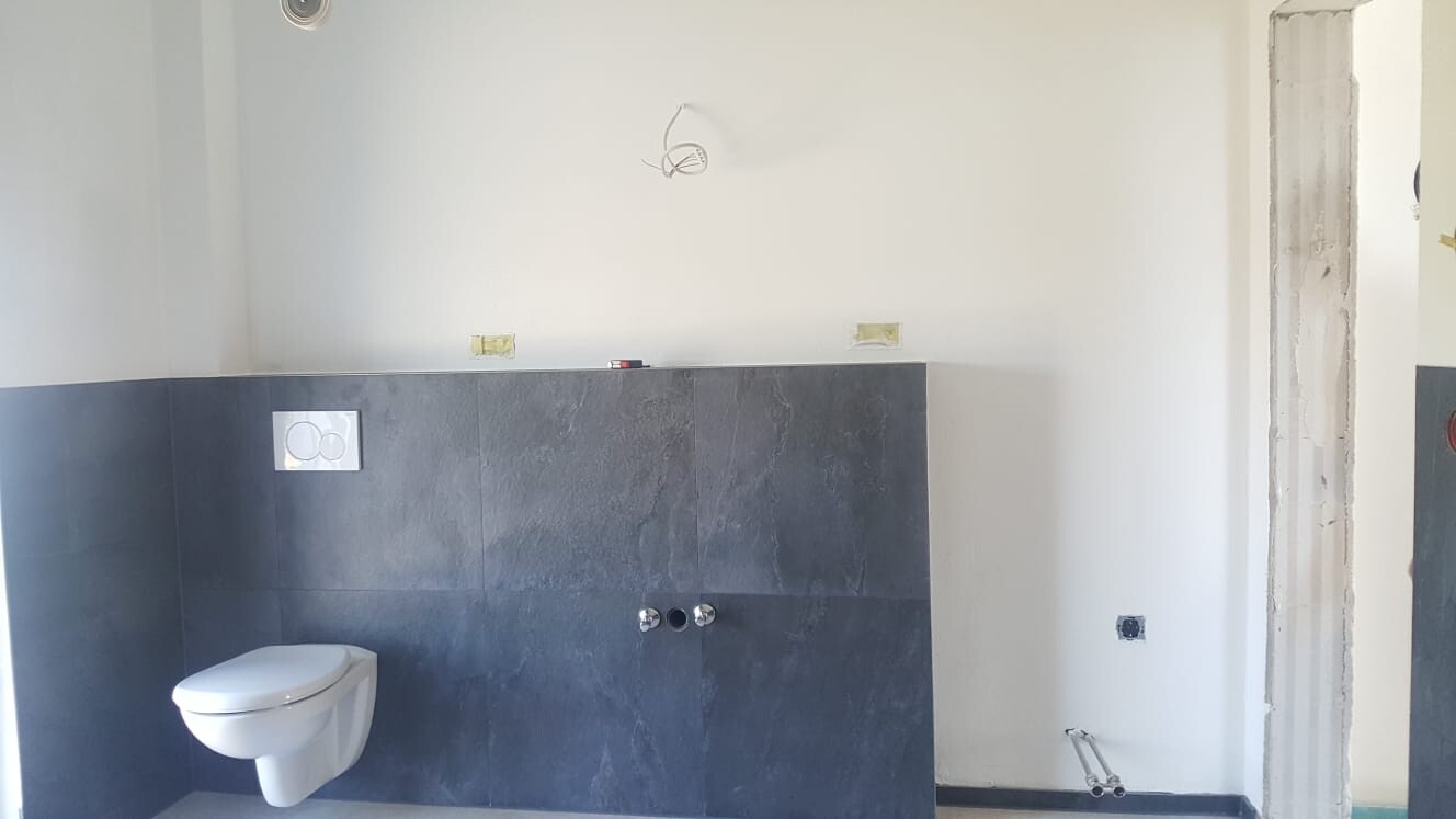 Badezimmer im Bau: dunkle Wandfliesen, wandhängendes WC links, offene Sanitärinstallationen