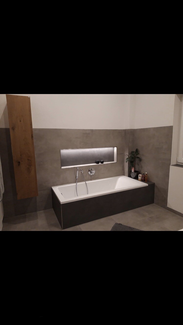 Moderne Badewanne in grauem Fliesenbad mit Wandablage, Pflanze und Badzubehör.