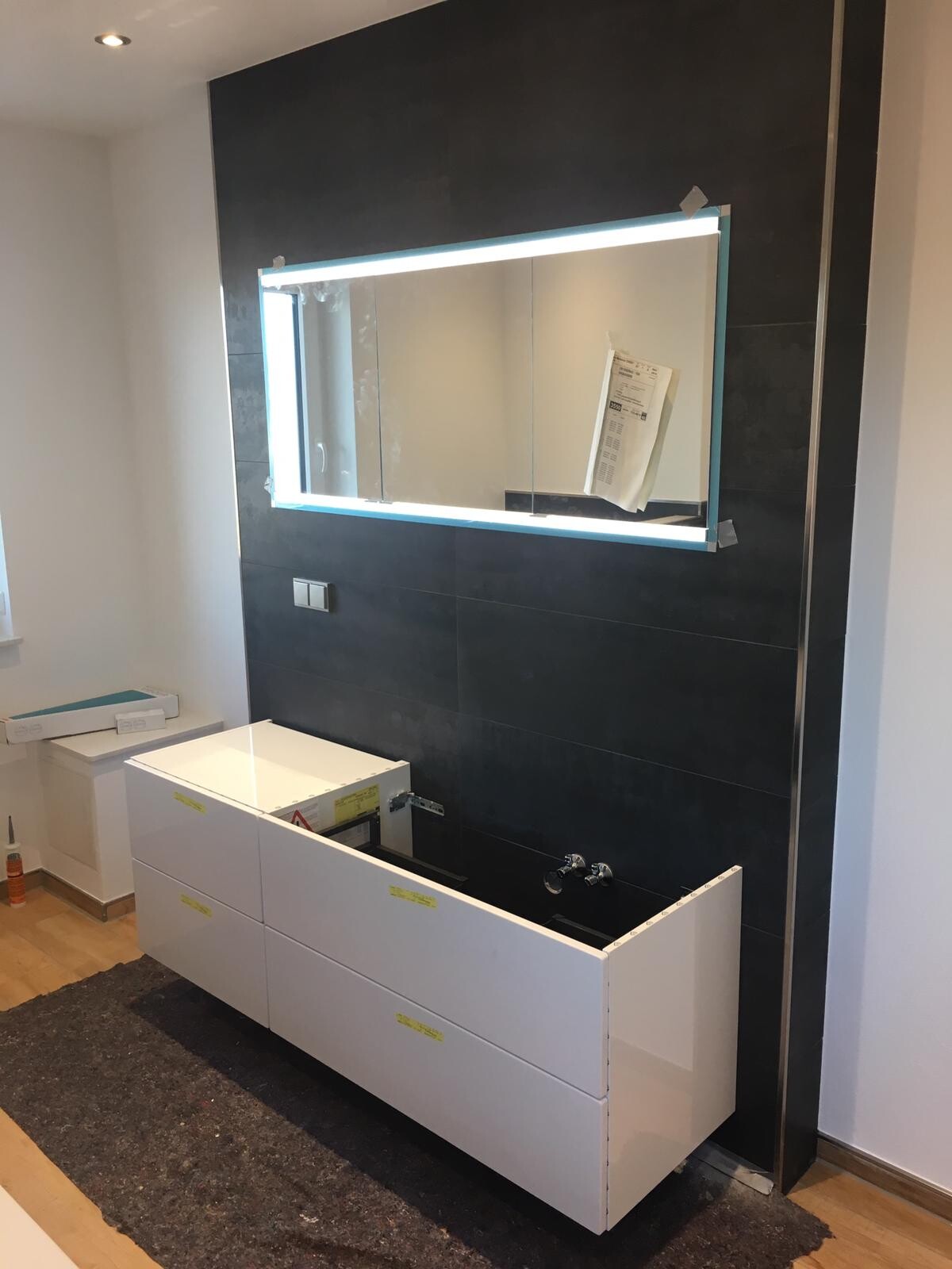 Modernes Badezimmer mit dunkler Fliesenwand, beleuchtetem Spiegel und unfertigem Waschtisch.