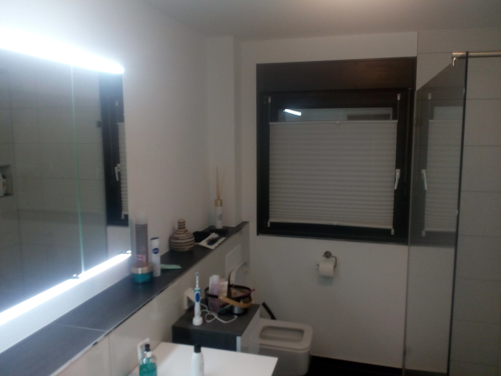 Modernes Badezimmer mit beleuchtetem Spiegel, Fenster mit Rollladen, Glasdusche und WC.