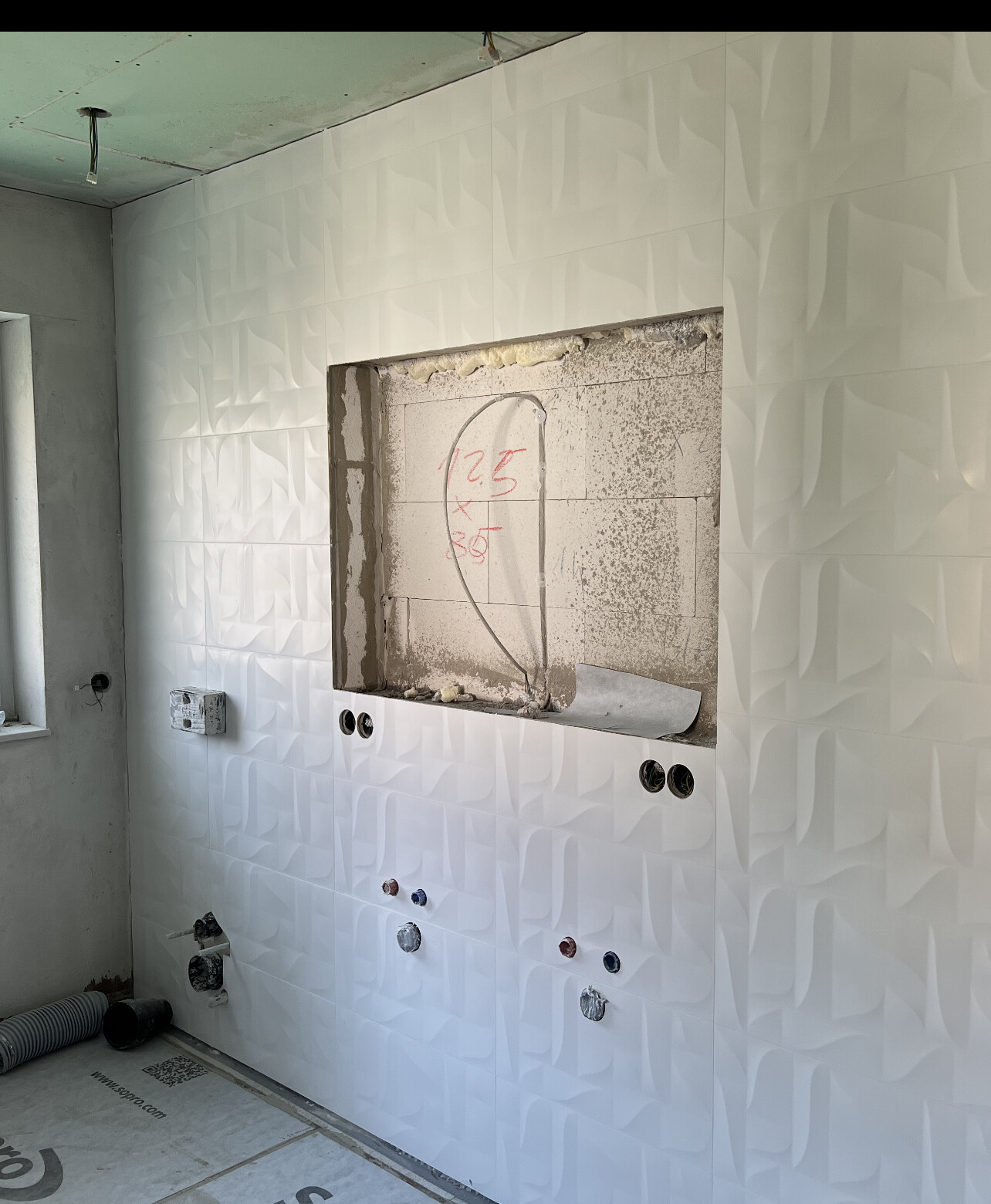 Badrenovierung: weiße 3D-Fliesen an Wand, Ausschnitt mit Rohbau und sichtbaren Rohren.