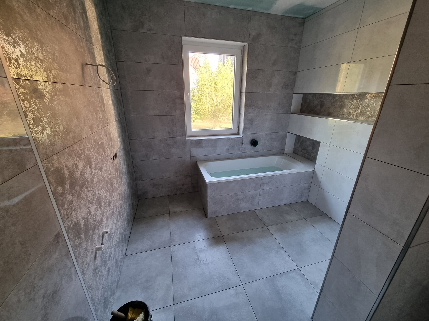 Badezimmer mit eingelassener Badewanne, grauen Fliesen und Fenster mit Blick nach draußen.