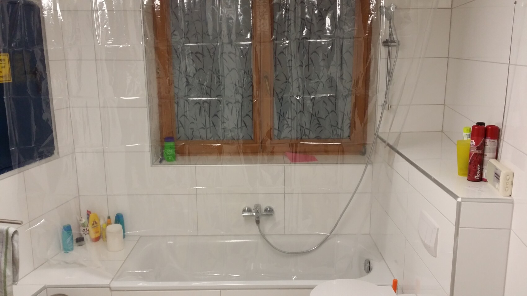Badezimmer mit Badewanne, Duschkopf, Fenster mit Musterfolie, Fliesen, mehrere Shampoos am Rand.