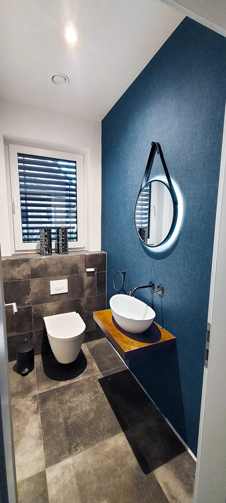 Modernes Bad: blaue Akzentwand, runder beleuchteter Spiegel, Waschbecken auf Holzplatte, WC.
