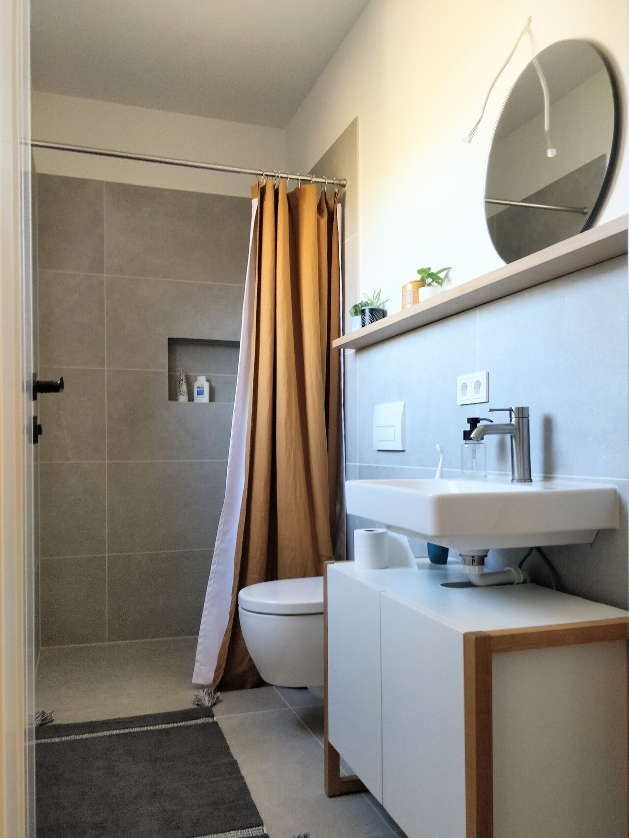 Badezimmer: Dusche mit braunem Vorhang, WC, Waschbecken, runder Spiegel, Pflanzen.