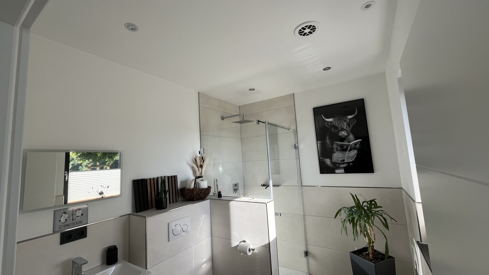 Modernes Badezimmer mit Glasdusche, Spiegel, Pflanze und Wandbild eines Stieres mit Zeitung