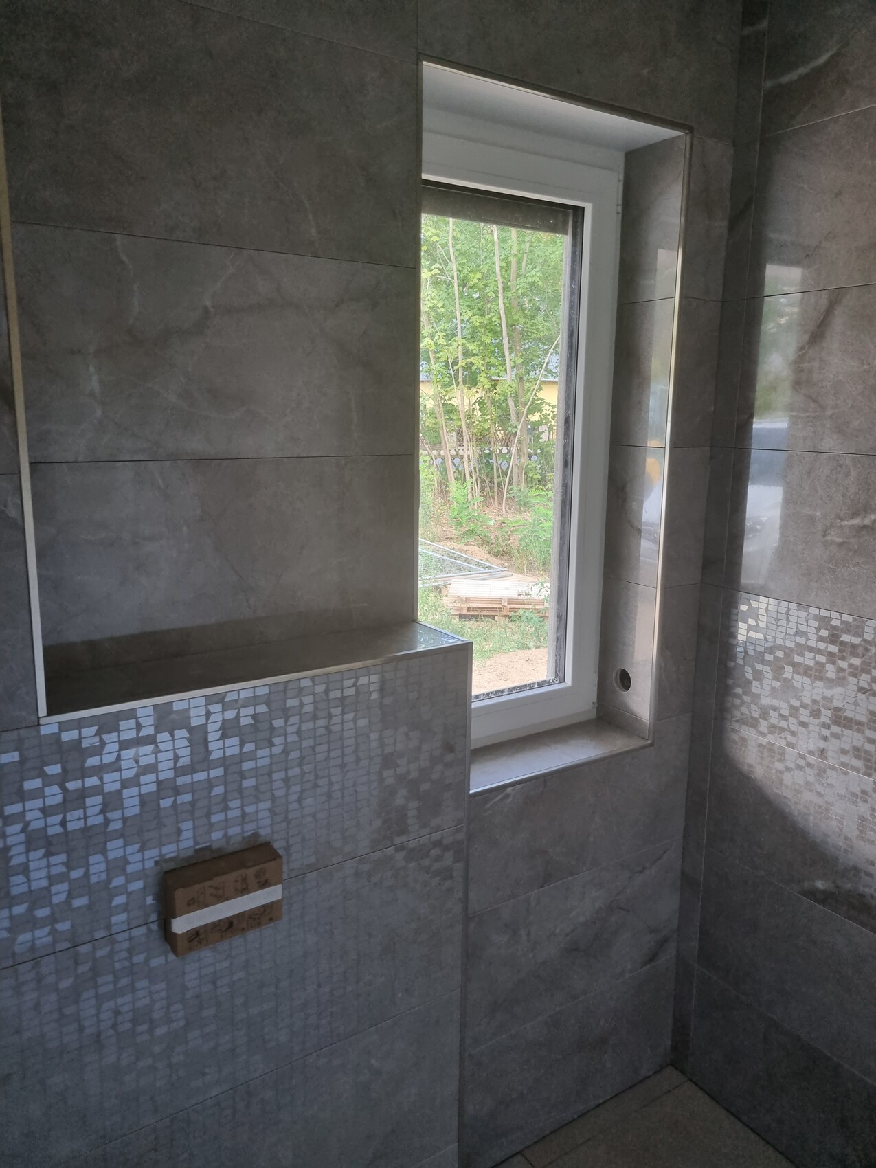 Badezimmer mit grauen Fliesen und Mosaikrand, weißes Fenster, Blick nach Garten.