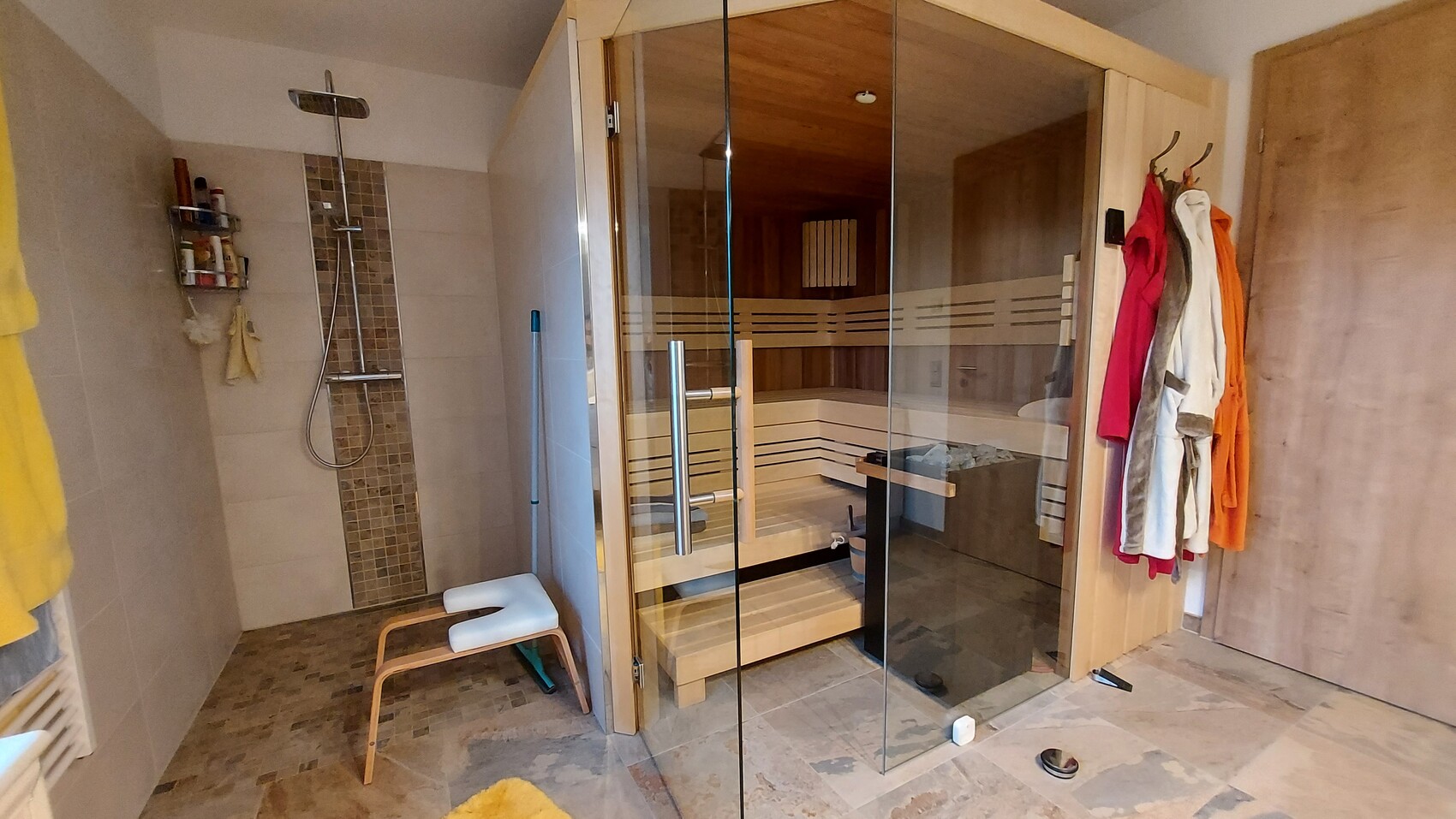 Badezimmer mit Regen-Dusche links, Glas-Sauna in der Mitte, bunte Bademäntel rechts.