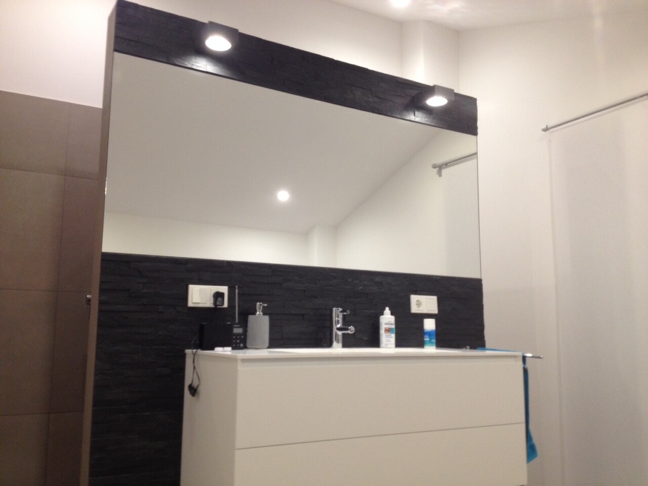 Modernes Badezimmer: weißer Waschtisch, großer Spiegel, schwarze Steinwand, blaues Handtuch rechts.
