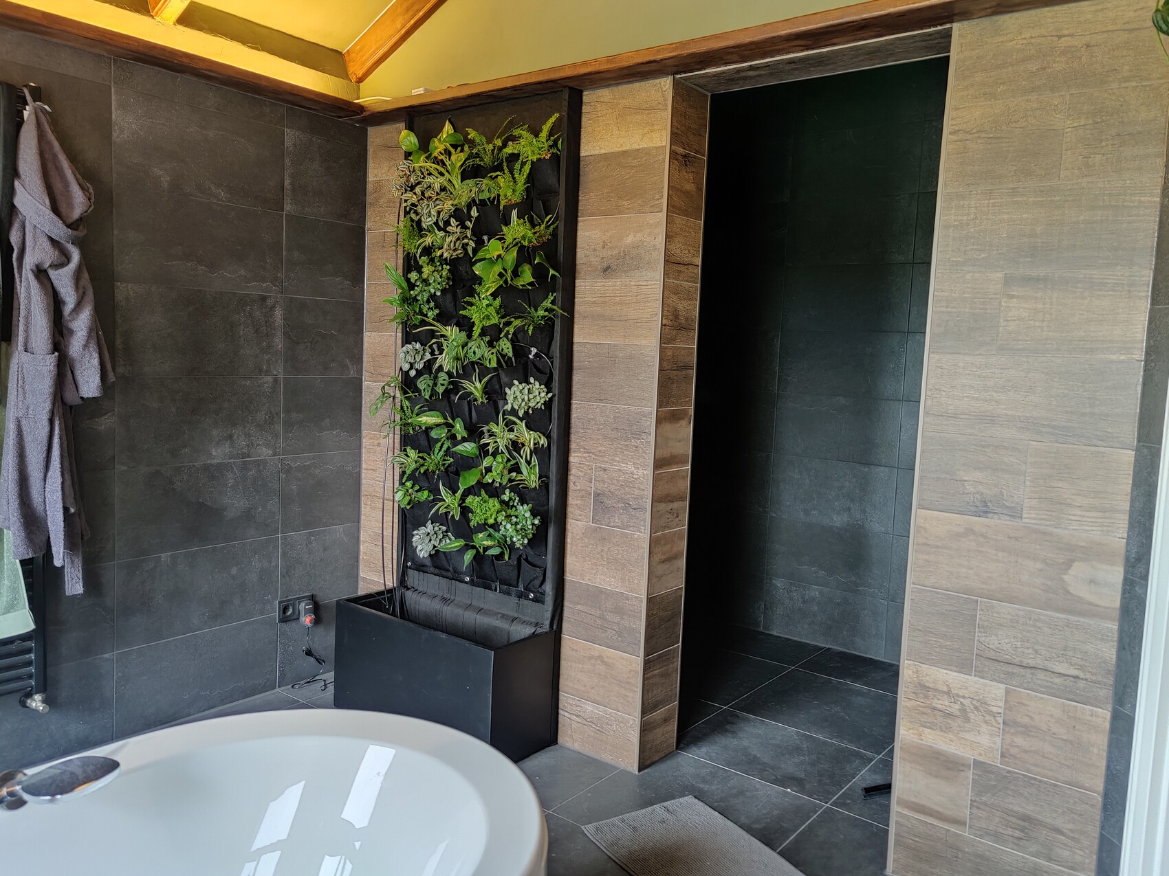 Modernes Badezimmer mit vertikaler Pflanzwand, dunklen Fliesen und einer freistehenden Badewanne.