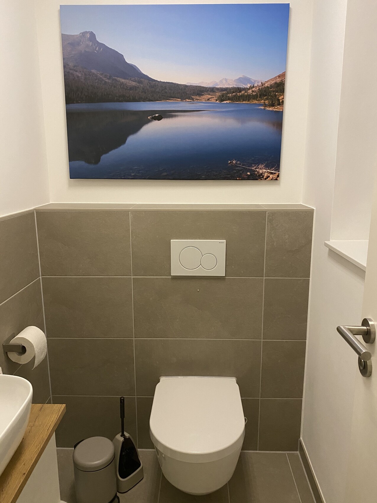 Badezimmer mit Toilette vor grauen Fliesen; darüber hängt Landschaftsbild eines Bergsees.