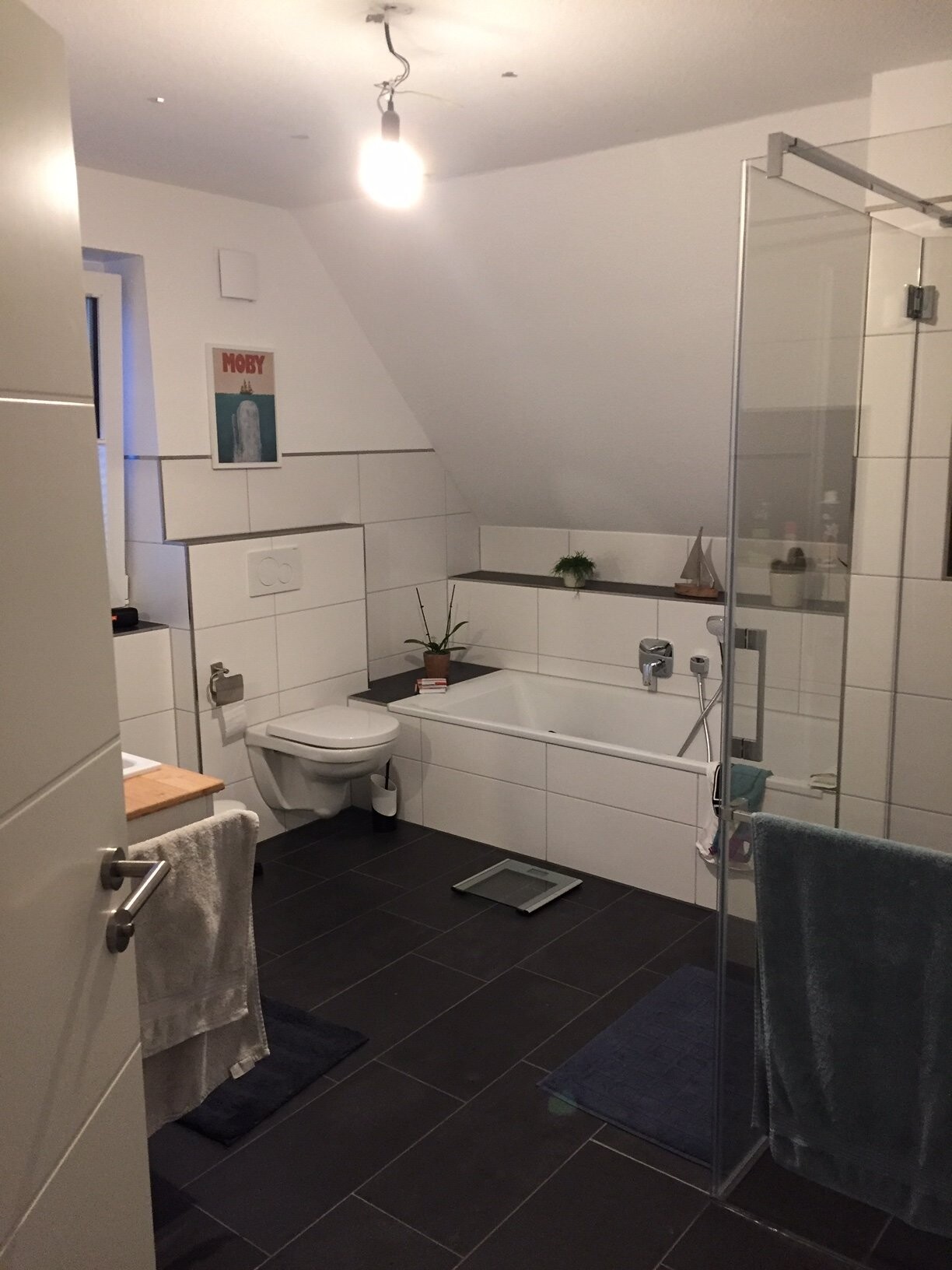 Helles Bad mit Badewanne, Glasdusche, WC, dunklen Bodenfliesen, Pflanzen und Poster.