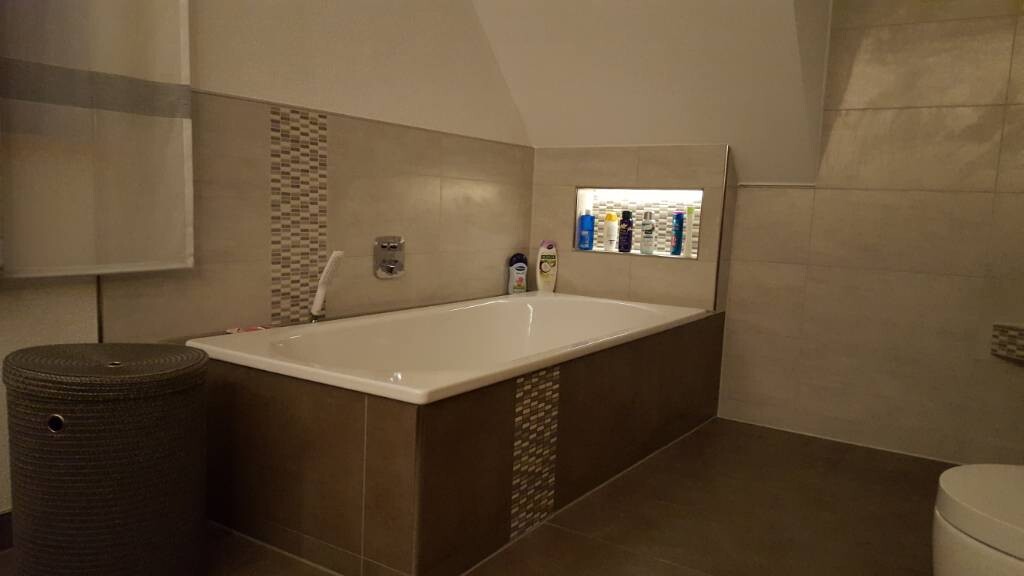 Moderner Badezimmerraum mit eingelassener Badewanne, braunen Fliesen und Ablagefach mit Pflegeprodukten