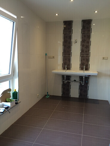 Badezimmer mit Waschbecken, dunklen Fliesen hinter dem Waschbereich und Fenster links