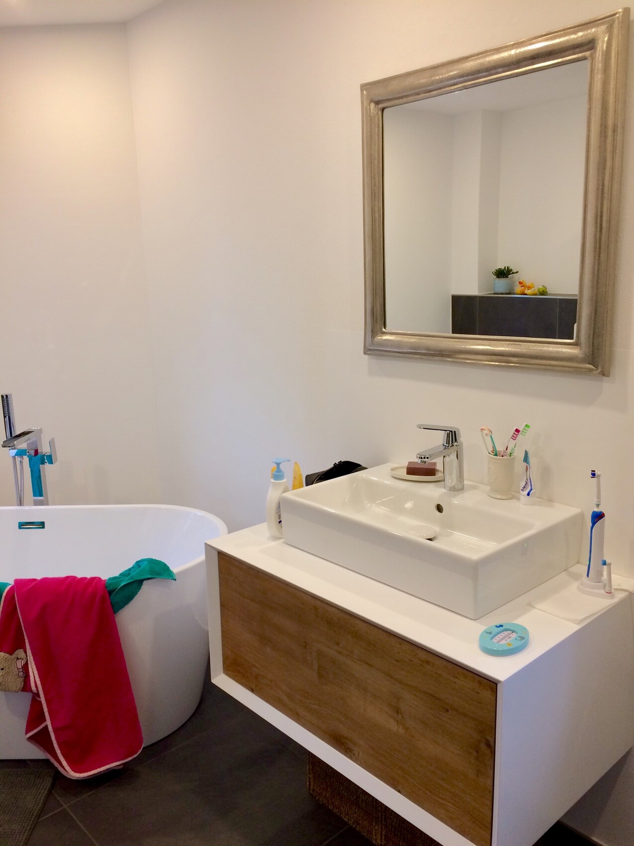 Modernes Badezimmer: Badewanne links, Waschbecken mit Holzunterbau, Spiegelrahmen, bunte Handtücher.
