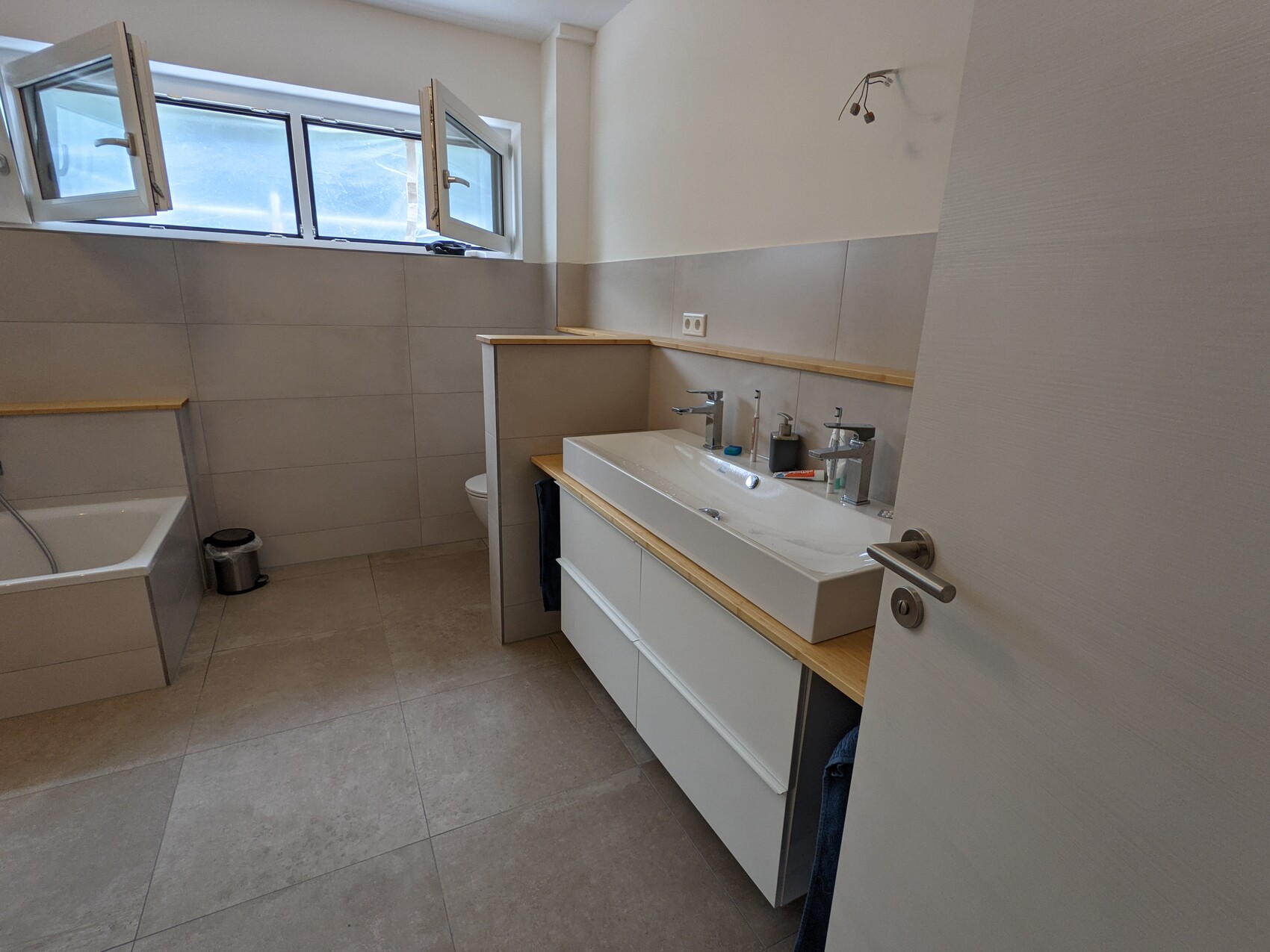 Badezimmer mit breitem Waschtisch, zwei Armaturen, Badewanne links, Fenster oben.