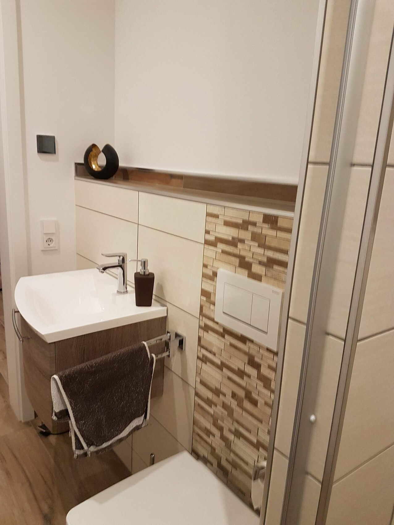 Modernes Badezimmer mit Waschbecken, Holzunterschrank, Mosaikwand, Handtuch, WC-Spülkasten und Dusche.