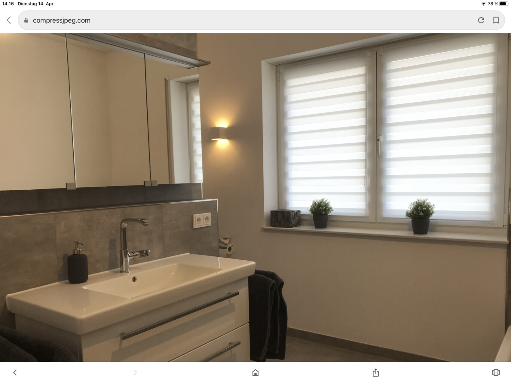 Badezimmer mit Waschbecken, grauer Fliesenrückwand, Fenster mit Rollläden, zwei Topfpflanzen.