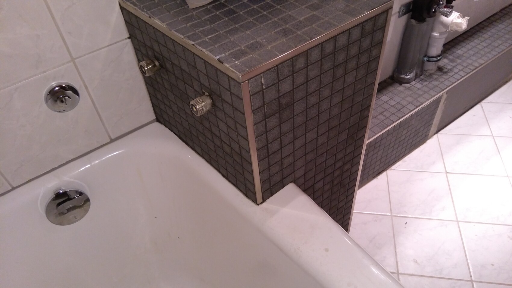 Eine Badewanne mit dunkler Fliesenverkleidung, seitlich sichtbare Wasseranschlüsse und Rohre.