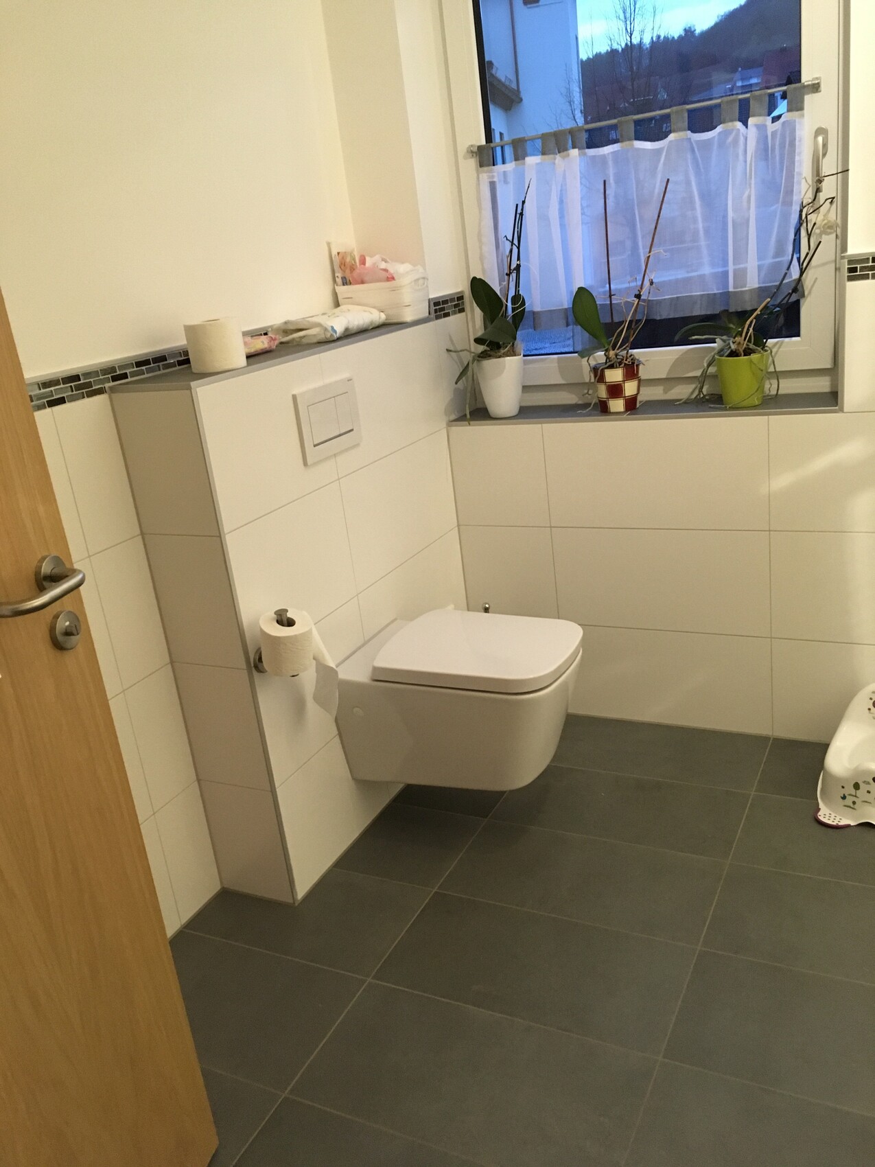 Modernes Badezimmer mit wandhängender Toilette, weißen Fliesen und Fensterpflanzen.