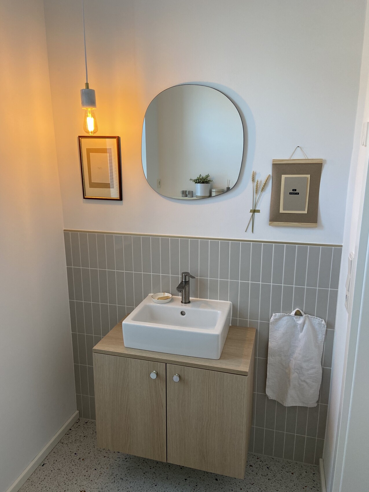 Kleines Badezimmer mit Holz-Unterschrank, Waschbecken, runder Spiegel, grauen Fliesen.
