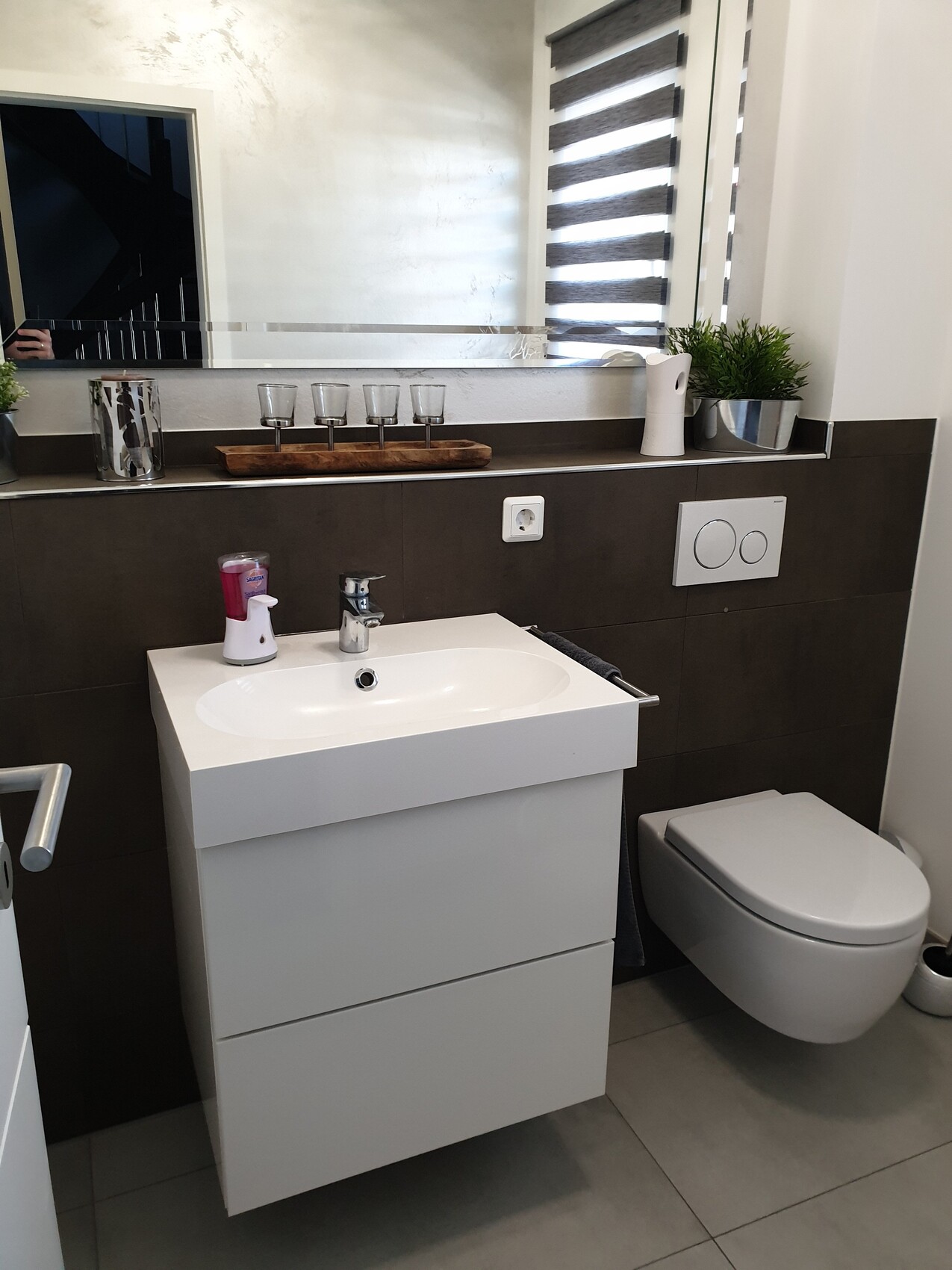 Modernes Bad mit weißem Waschtisch, wandhängende Toilette, Spiegel, Fensterjalousie, Pflanze.