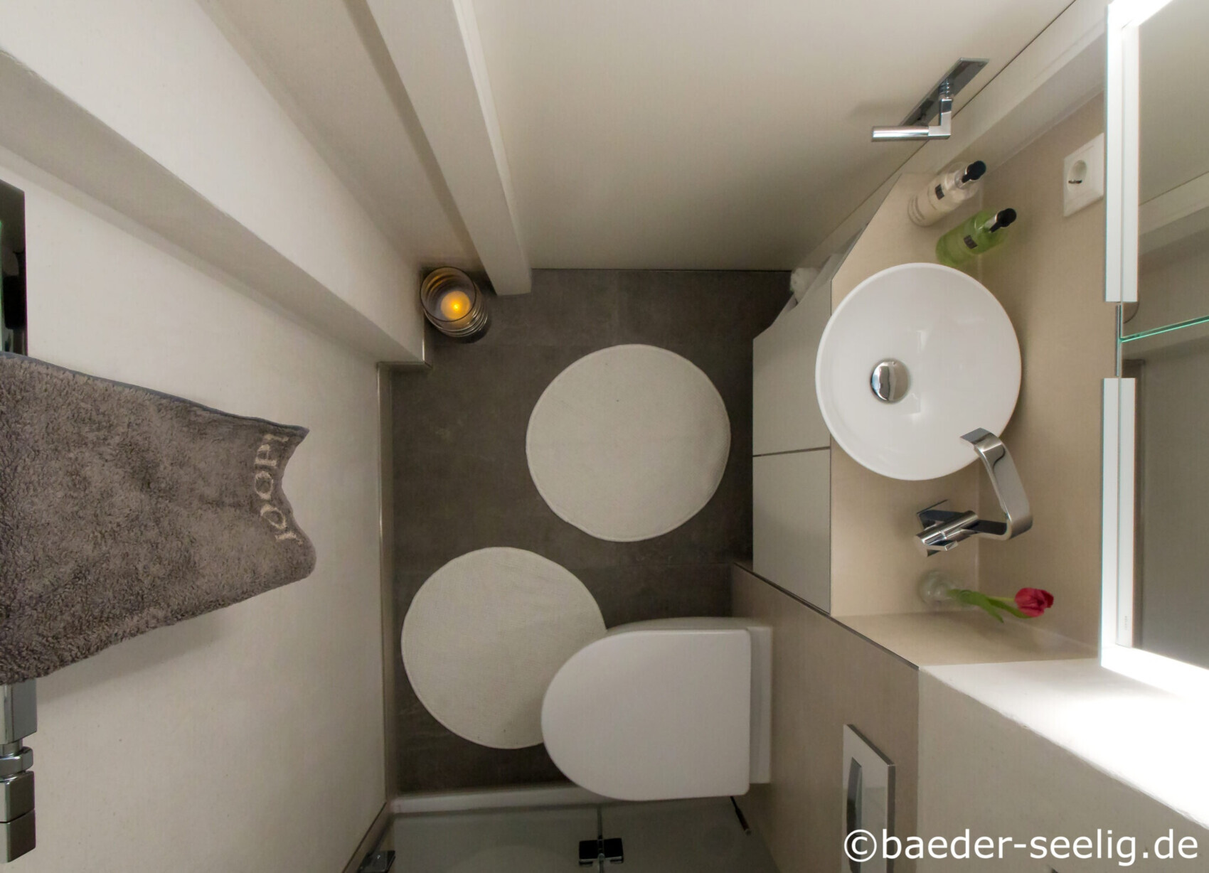 Kleines Bad von oben: runder Waschbecken, Toilette, zwei runde Teppiche, Handtuch an der Wand.