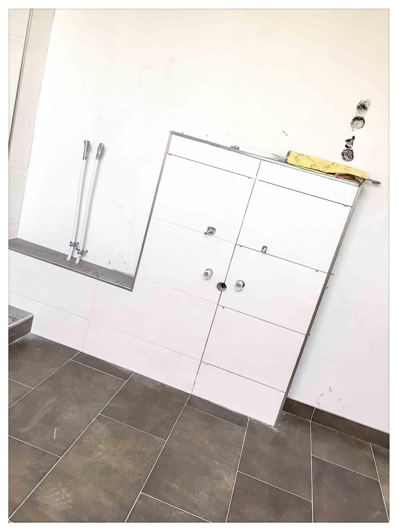 Weißer Badezimmerschrank lehnt schräg an der Wand, sichtbare Rohre, dunkle Bodenfliesen.