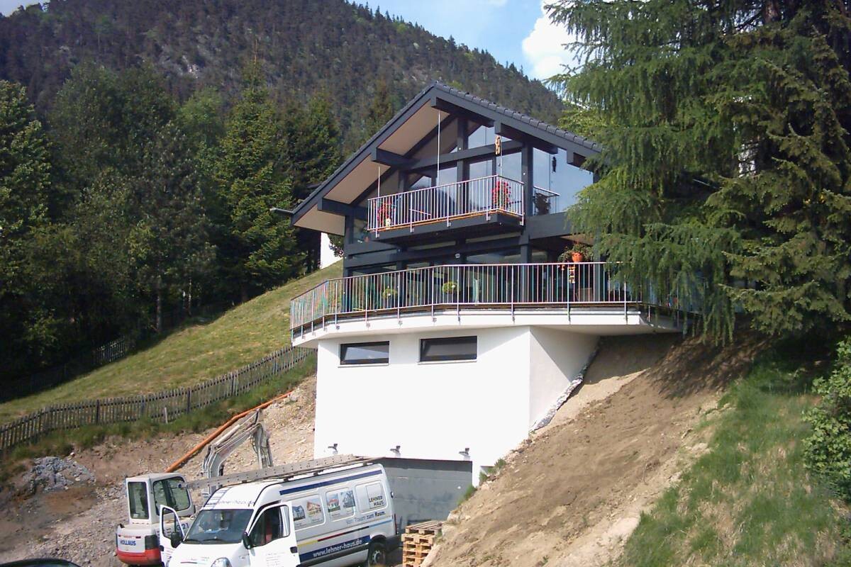 Modernes Chalet auf Hang mit Glasfront und umlaufender Terrasse; Baumaschinen unten.