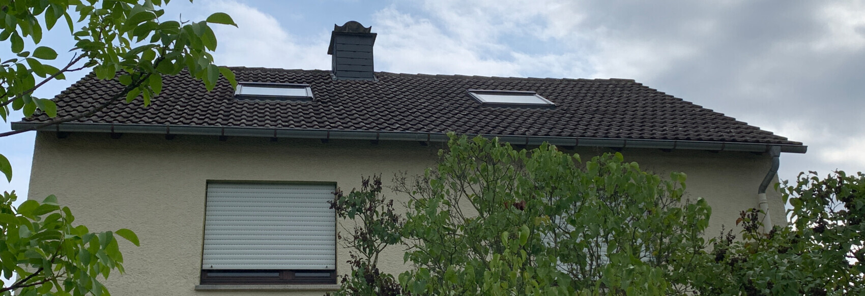 Zweistöckiges Haus mit braunen Ziegeln, zwei Dachfenstern, Schornstein und Bäumen davor