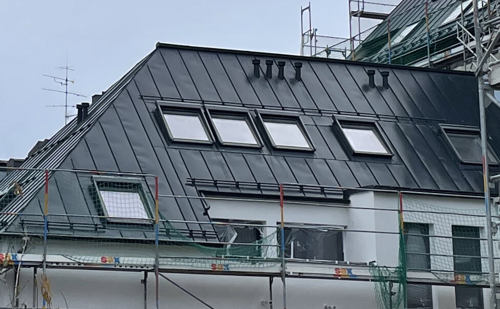Schräges, dunkles Metalldach mit drei Dachflächenfenstern; Baugerüst und Netze an der Baustelle.