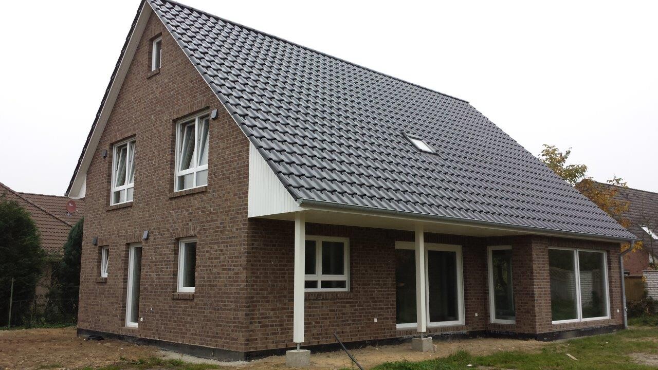 Modernes braunes Backsteinhaus mit steilem grauen Ziegeldach, großen Fenstern und Veranda.