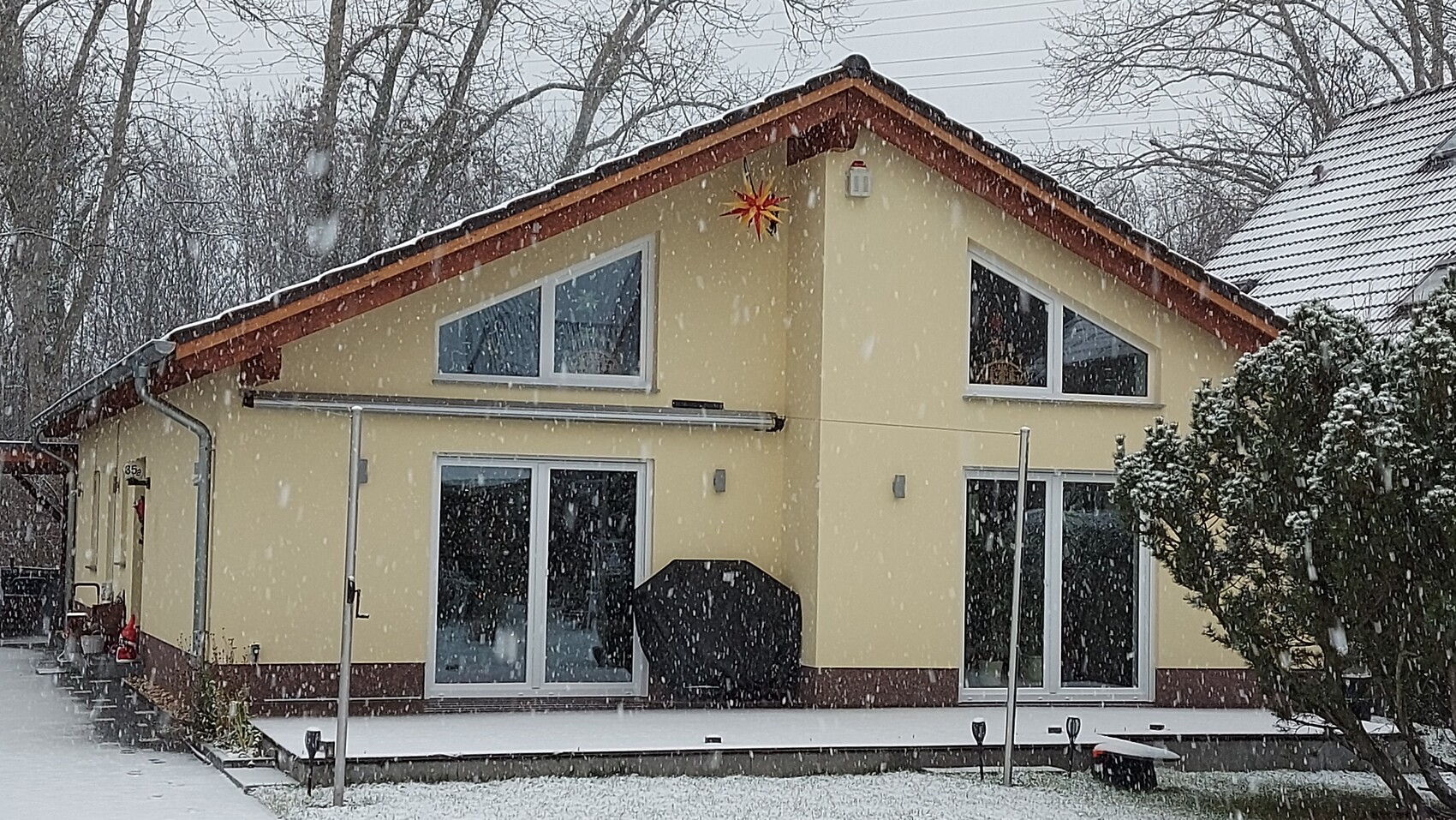 Gelbes Einfamilienhaus mit großen Glasfronten im Schneefall, Terrasse mit Grill