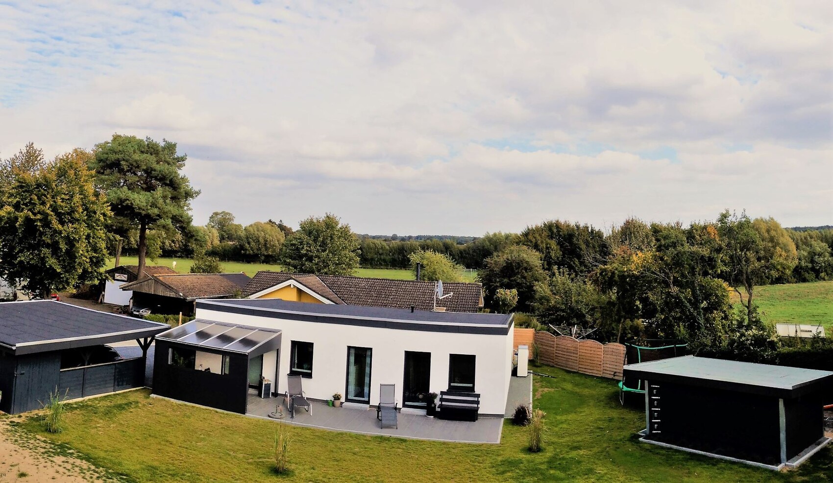 Moderne weiße Villa mit Terrasse, Garten, Trampolin und ländlicher Landschaft.