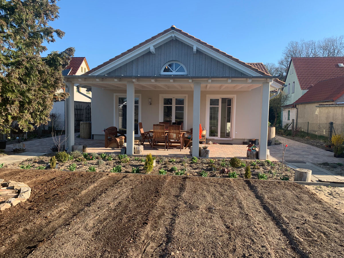 Überdachte Terrasse vor modernem Haus mit Esstisch und Stühlen; Gartenbeet im Vordergrund.
