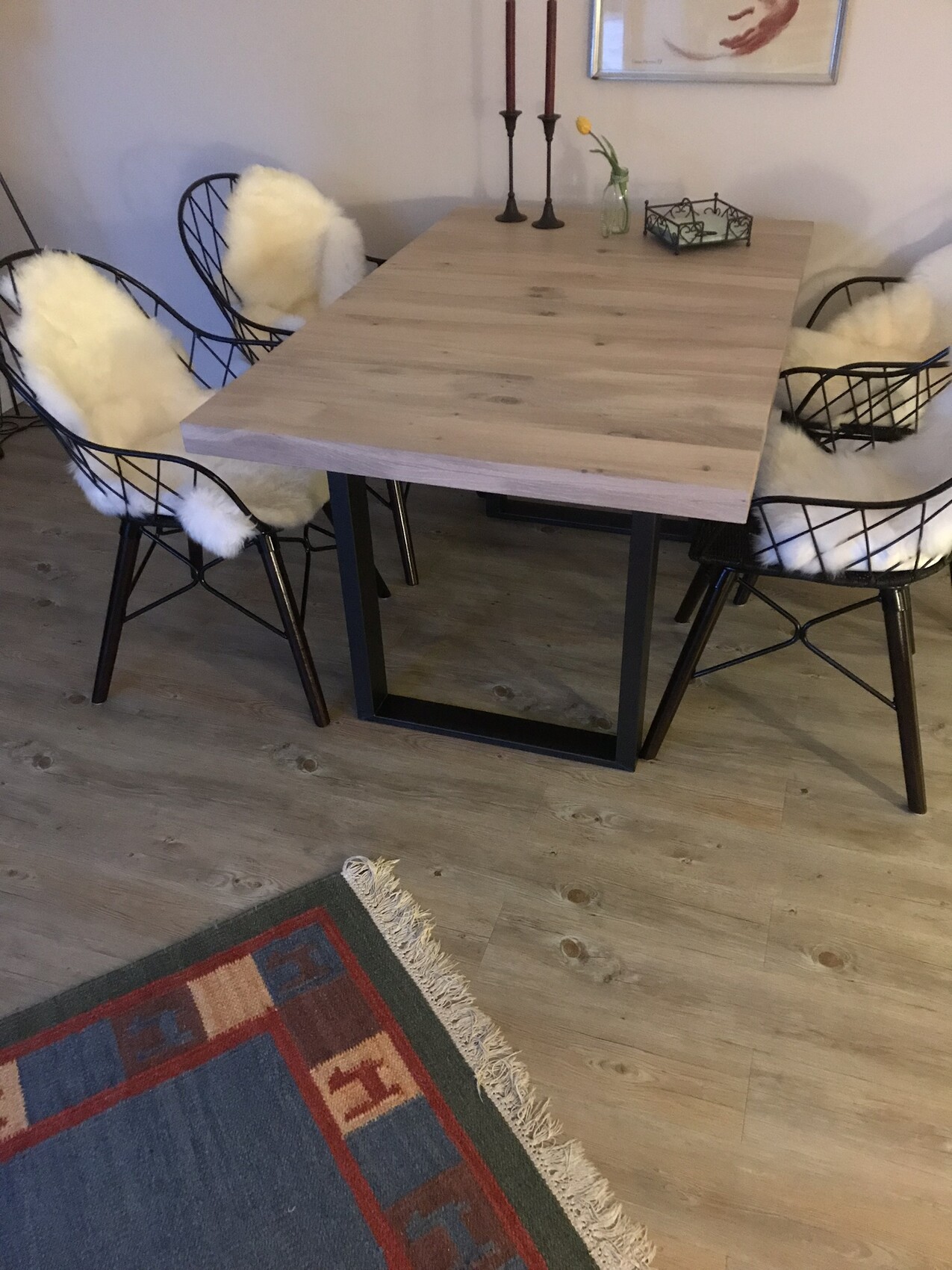 Esstisch aus hellem Holz mit schwarzen Metallbeinen, vier Stühle mit weißen Fellkissen, Kerzenständer und Vase.
