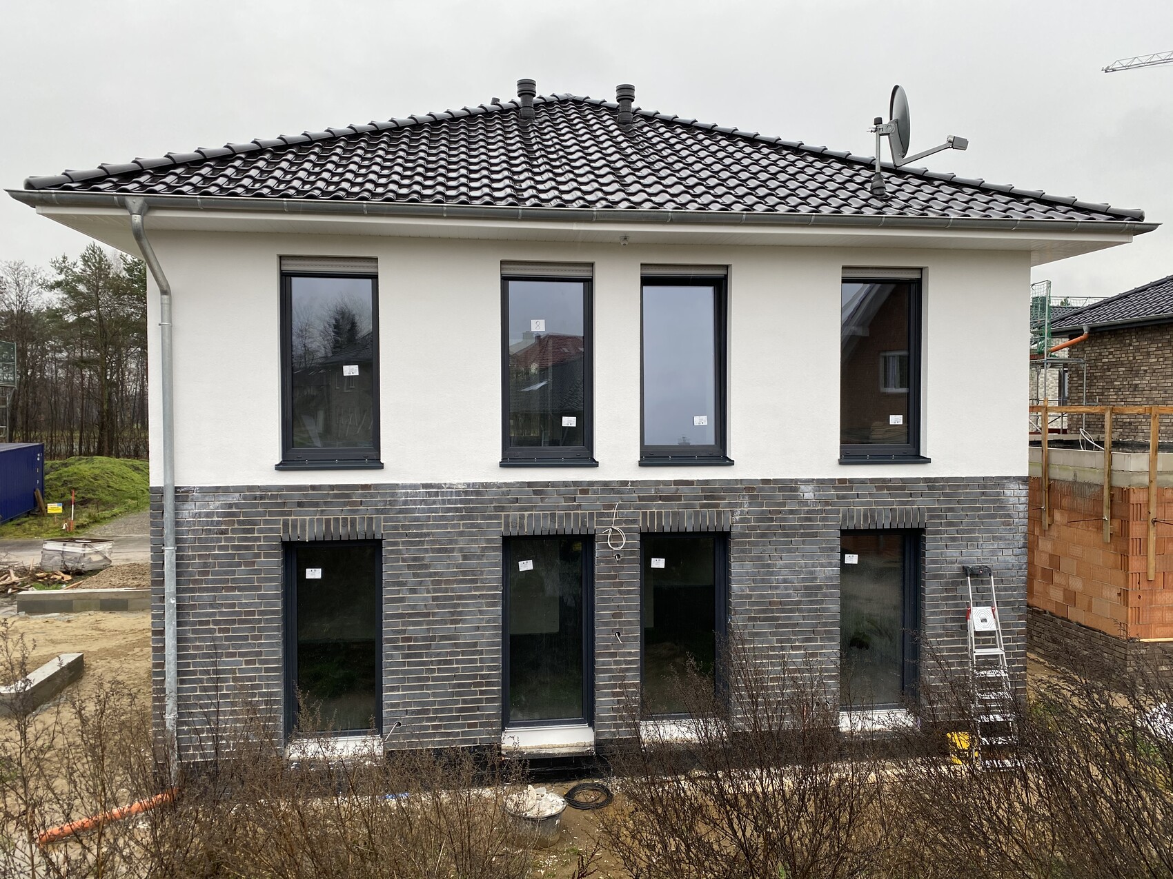 Zweistöckiges Neubauhaus; weiße Oberseite, dunkler Ziegelunterbau, bodentiefe Fenster; Leiter rechts.