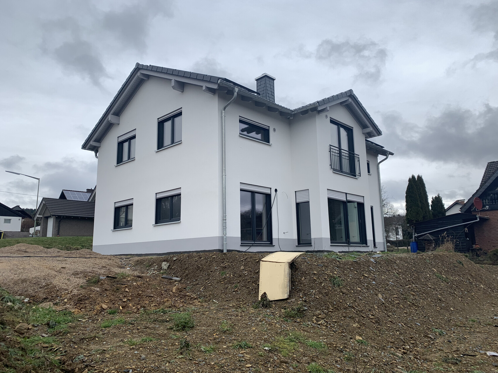 Weißes, modernes zweistöckiges Haus mit Balkon, großen Fenstern und grauem Dach; Baugrund davor.