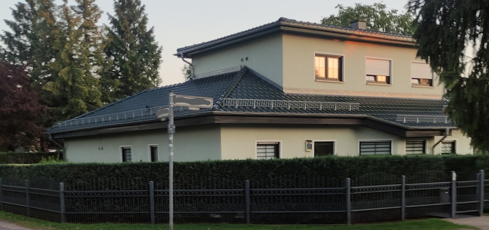 Zweistöckiges Haus mit blauem Ziegel-Dach, von einer Hecke und einem Metallzaun umgeben.