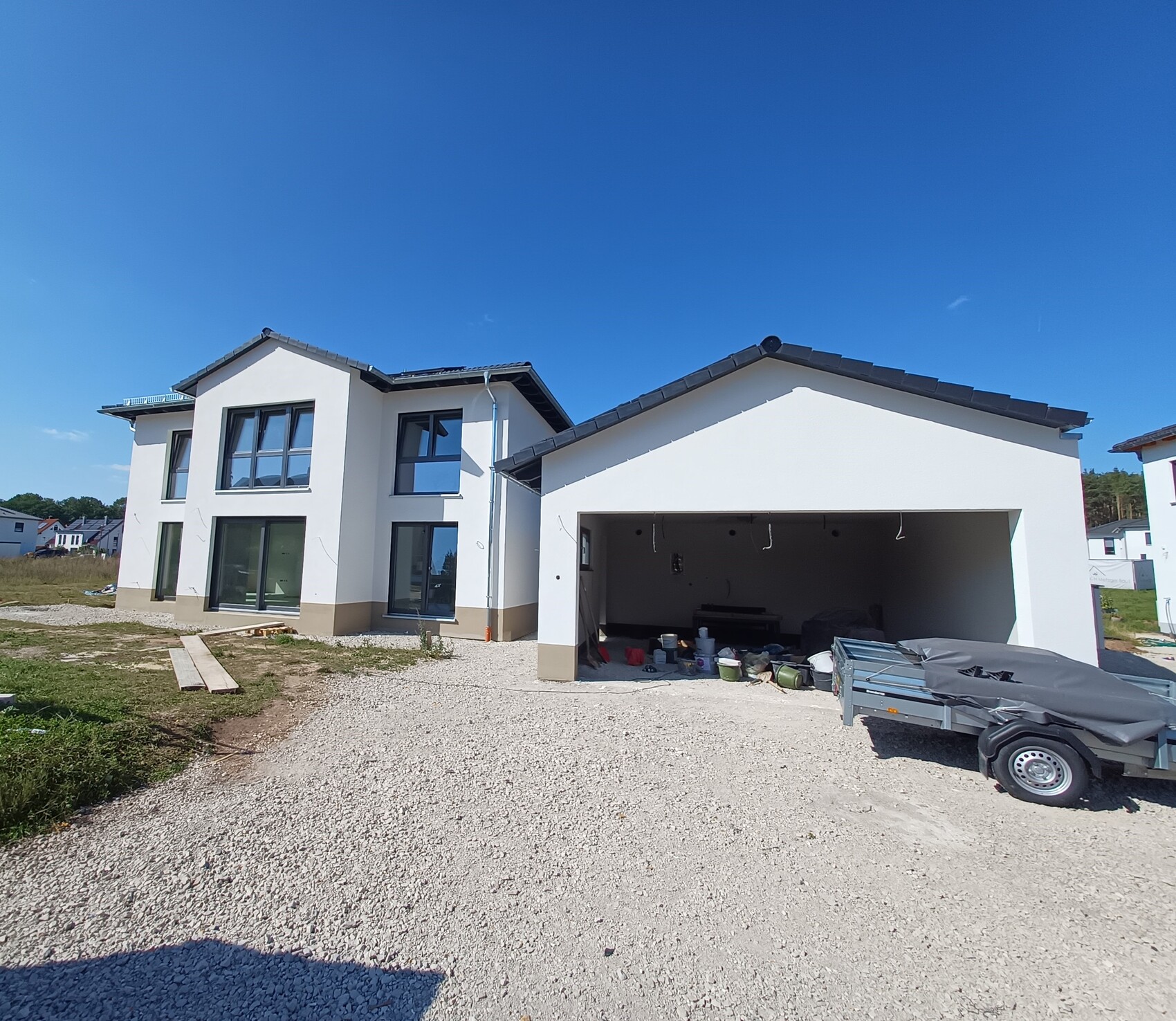 Modernes weißes Einfamilienhaus mit offener Garage, Bauutensilien und Anhänger davor.