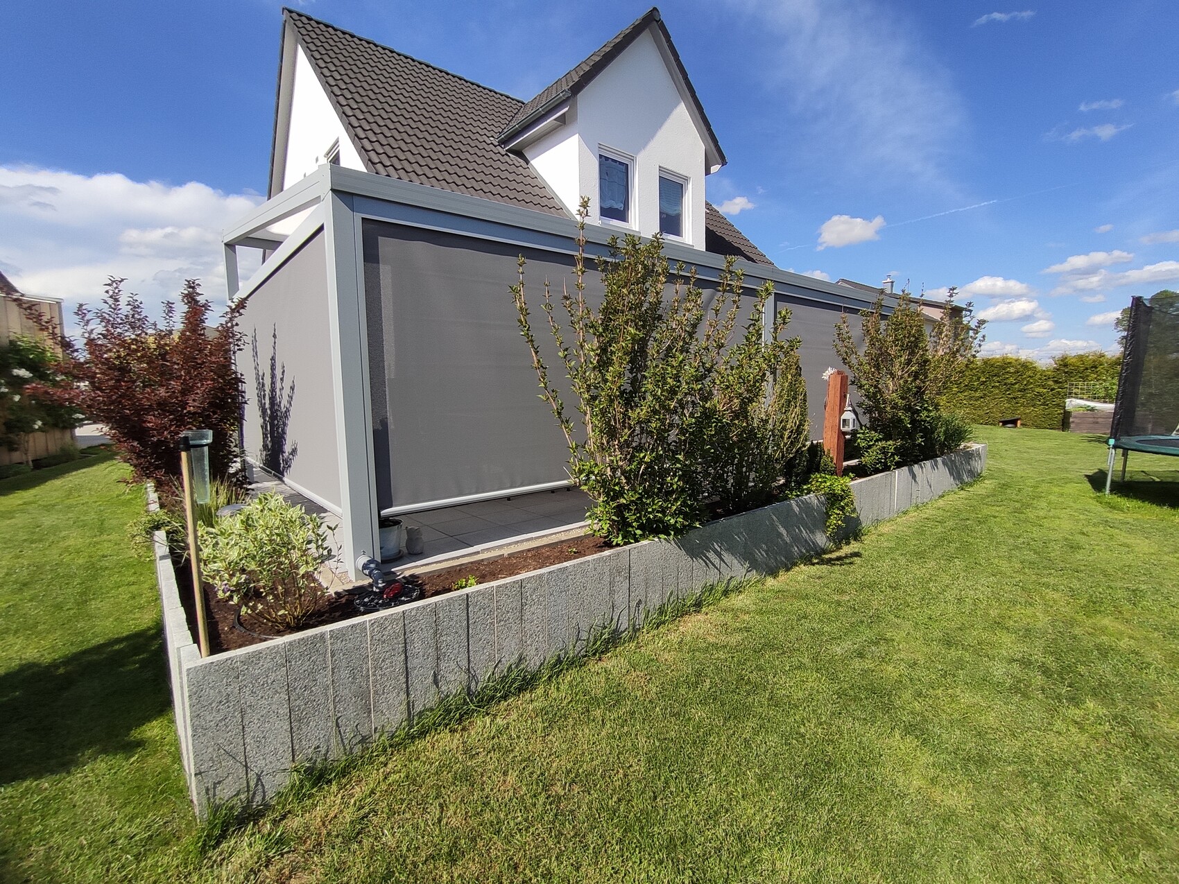 Zweistöckiges weißes Haus mit grauem Anbau, erhöhte Betonbeete, Garten mit Bäumen und Trampolin.