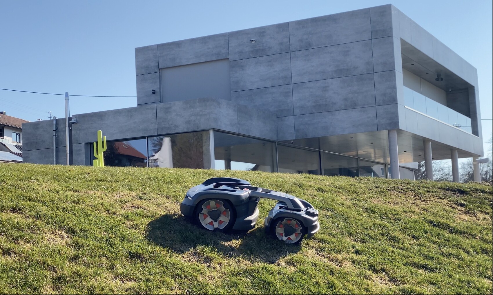 Modernes graues Betongebäude mit Glasfront, grüner Kaktus-Skulptur, Spielzeugauto im Gras.