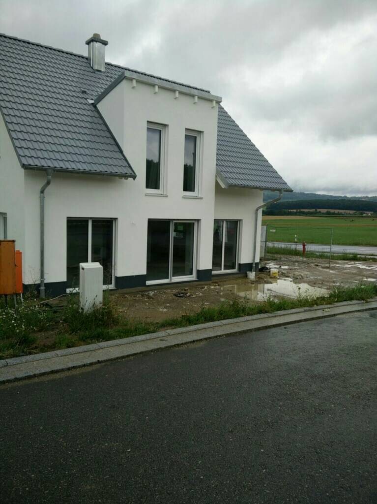 Weiße moderne Einfamilienhausfront mit großen Glasfenstern, Bauplatz davor, bewölkter Himmel